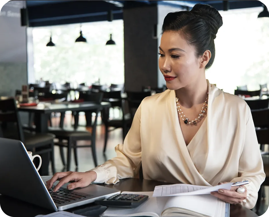 Multi-unit restaurant operator reviewing a printed financial report at a laptop in an empty dining room, with a calculator and ledger on the table