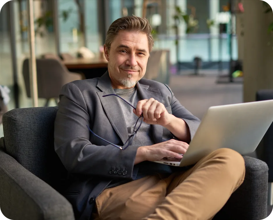 A man in a grey jacket sitting back in an armchair in a modern office lounge, holding his glasses in one hand and working on a laptop balanced on his knee