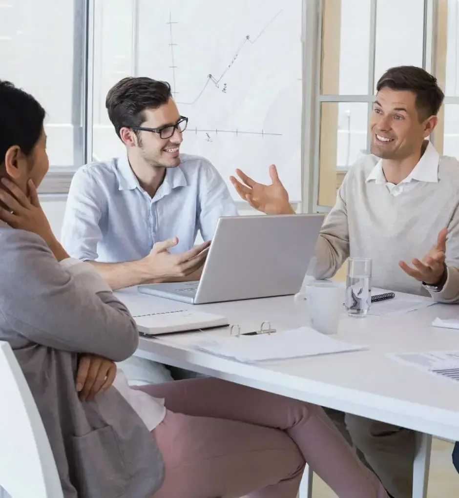 Three people talking around an open laptop at a white meeting table, with a hand-drawn growth chart on a whiteboard behind them and papers, glasses and a notebook spread across the table
