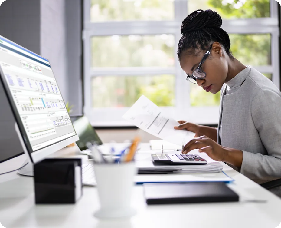 Property accountant at a desk reviewing a printed statement against a financial system on screen, with a calculator and files alongside