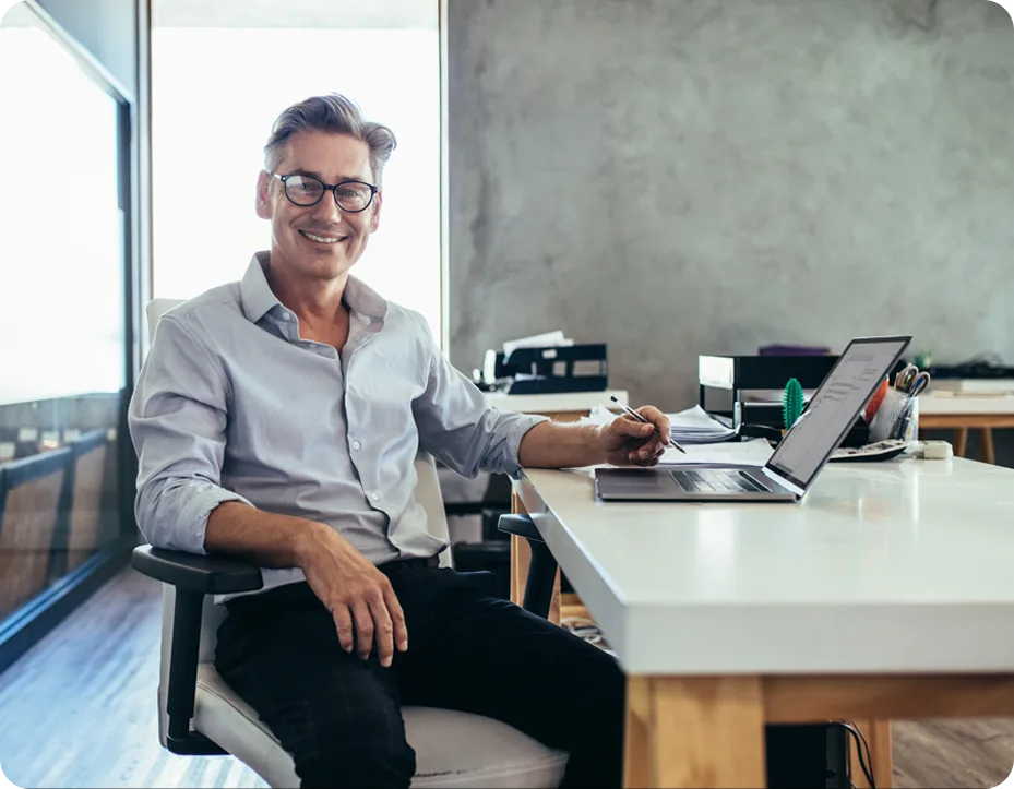 Man in a grey shirt and dark-framed glasses seated at a white desk in a modern office, an open laptop and printed notes in front of him and a floor-to-ceiling window behind