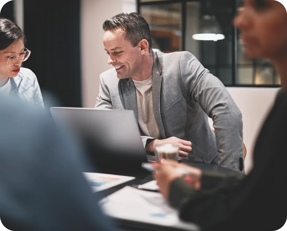 Three colleagues in a dark modern office leaning over an open laptop together, one of them smiling, with printed charts and a coffee glass on the table in front of them